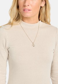 Top beige a maniche lunghe con righe, scollatura alta e una texture sottile. Collana dorata con ciondolo circolare che esalta la scollatura.