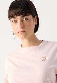 Felpa a maniche corte, di colore rosa chiaro, con collo rotondo e un piccolo logo geometrico marrone sul lato sinistro del petto. Tessuto di cotone liscio.