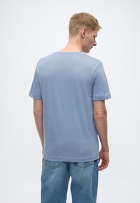 Šviesiai mėlynas trumpomis rankovėmis marškinėlis, pagamintas iš minkštos, lygaus audinio, su klasikine apvalia iškirpte ir laisvu siluetu, dėvimas su išblukusiais džinsais.