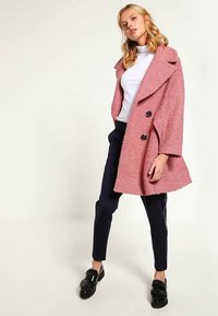 Donna che indossa un cappotto doppiopetto rosa texturizzato, un dolcevita bianco, pantaloni neri e mocassini neri, in piedi con una mano in tasca del cappotto.