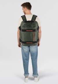 Mochila verde con correas ajustables, acentos negros y detalles naranjas. Presenta un bolsillo delantero con cremallera y un tejido texturizado y duradero.