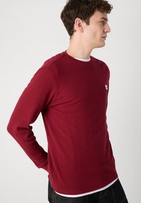 Bordeauxkleurig langarmshirt van een textuurstof met een geribde structuur, ronde hals en een klein etiket op de borst. Draagt over een wit shirt.