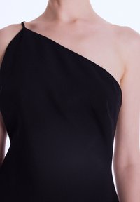 Femme portant un haut sans manches noir à une épaule, découvrant son cou, son épaule et le haut de sa poitrine sur un fond uni.