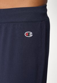 Marinblå bomulls joggebukser med ribbestrikket liv og en liten brodert logo i hvitt, rødt og blått på venstre lår. Myk tekstur.