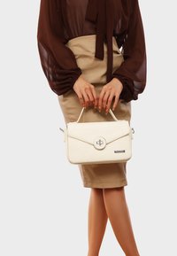 Borsa bianca con texture, dotata di patta, hardware circolare argentato e manico corto, abbinata a una blusa marrone e una gonna beige.