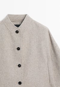 Lichtbeige wollen jas met een staande kraag en vier donkere knopen aan de voorkant, getoond op een witte achtergrond.