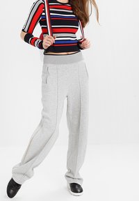 Graue Jogginghose mit seitlichen Reißverschlüssen, strukturiertem Stoff und elastischem Bund. Kombiniert mit einem schwarzen, roten und blauen gestreiften Crop-Top und Trägern.