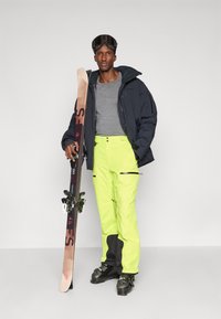 Mies neonkeltaisissa hiihtohousuissa, mustassa hiihtotakissa ja suojalaseissa pitää hiihtosuksia ja seisoo vaaleaa taustaa vasten.