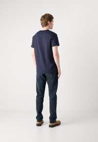 T-shirt à manches courtes bleu marine associé à un jean bleu foncé et des chaussures marron clair, vu de dos sur un fond blanc uni.