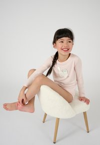 Un haut de pyjama à manches longues rose avec un graphique de chat, associé à un ton de peau clair. L'enfant est assis sur une chaise blanche fluffy avec des pieds en bois.