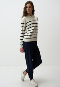 Pull à capuche tricoté en crème avec des rayures bleu marine, associé à un pantalon de jogging bleu marine et des baskets blanches, mettant en avant une coupe décontractée et un design casual.