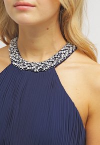 Marinblå plisserad klänning med en glittrande krystalliknande dekoration runt halsen, som visar en halterneck-design och slät textur.