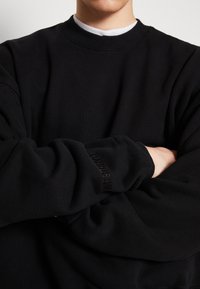 Černá mikina s kulatým výstřihem a ribbenými manžetami. Má měkkou, strukturovanou tkaninu a jemný vyšitý detail na rukávu.