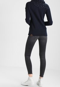 Kvinna i mörka smala jeans, vita högstövel sneakers och en marinblå hoodie med mönstrad huva, sedd bakifrån mot en vit bakgrund.