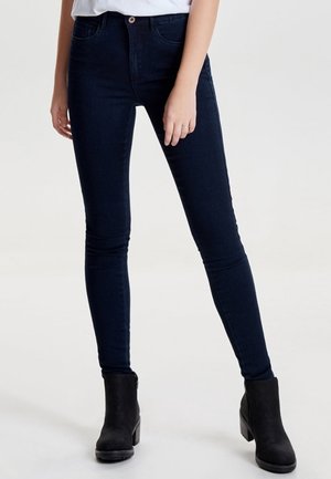 Personne portant un jean skinny bleu foncé, des bottines noires à talons blocs et un haut blanc, debout devant un fond clair uni.