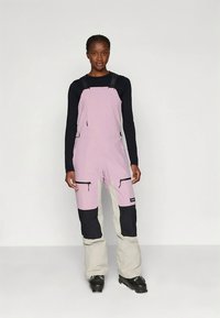 Rosa og svarte skibukser med justerbare stropper, glidelåslommer foran og et kontrasterende kamelfarget nedre parti, med en avslappet passform og slitesterkt stoff.