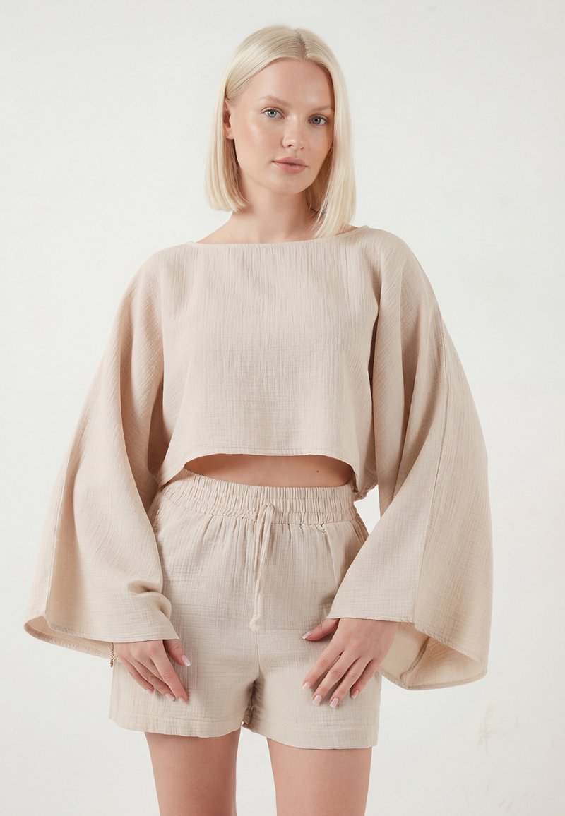 Mujer rubia con top corto de manga larga suelta y texturizada en color beige, y pantalones cortos a juego de cintura alta, posando contra un fondo liso.