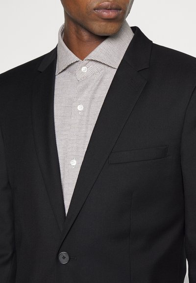 Schwarzer schmal geschnittener Blazer mit einem einzelnen Knopf, kombiniert mit einem strukturierten grauen Hemd, das ein kleines, dezentes Muster und weiße Knöpfe aufweist.