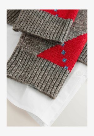 Pelēks adīts džemperis ar ribotu piedurkņu malām, kuram ir sarkanas trīsstūrveida formas raksti un zili punkti; uzlikts uz baltas krekliņas ar apkakli.