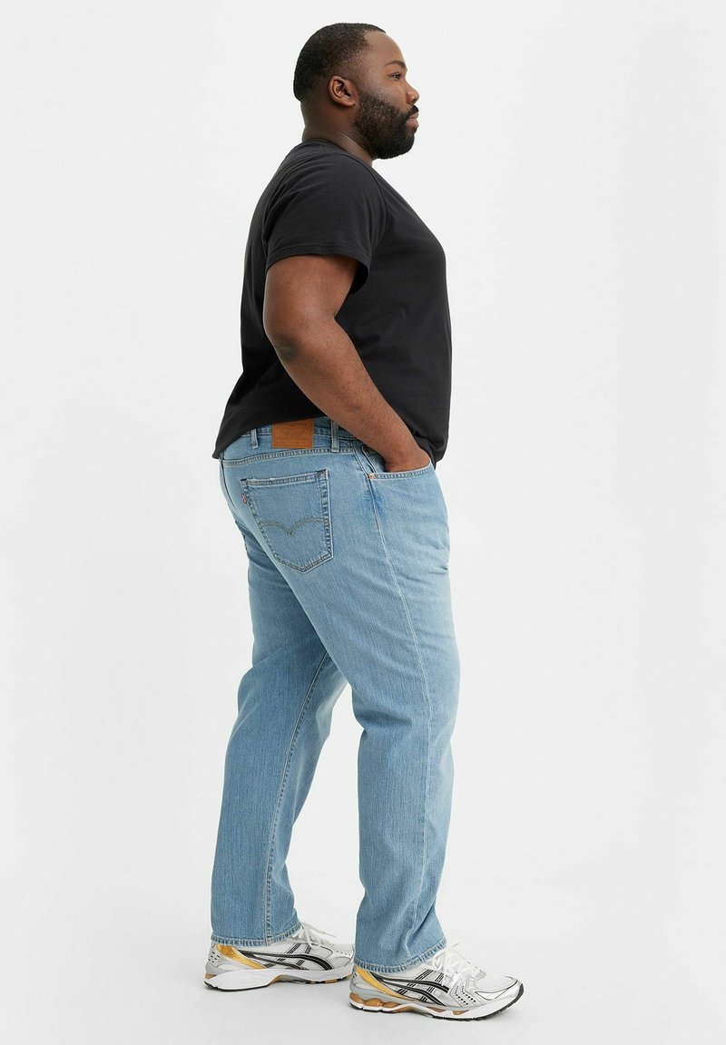 Helles blaues Denim mit geradem Bein, ausgestattet mit Taschen und einem braunen Lederpatch; getragen mit einem schwarzen T-Shirt und weißen Sneakern.