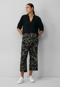Blouse noire à manches courtes avec un col en V et un tissu texturé, associée à un pantalon large cropped à motifs feuilles noirs et crème et des baskets blanches.