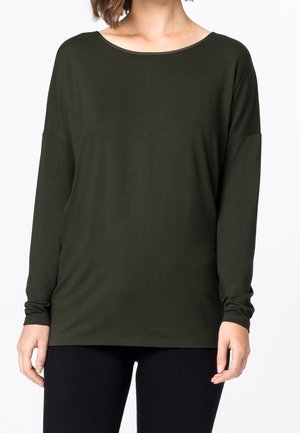 Woman wearing a long-sleeve, dark green loose top with a wide round neckline and black fitted pants against a white background.