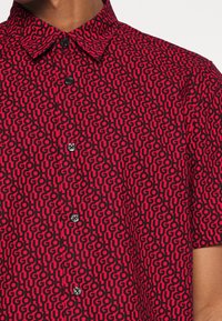 Camisa de manga corta con un vibrante patrón rojo y negro que presenta letras estilizadas. Cuello con botones y botones en contraste en la parte delantera.