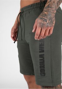 Olivengroene shorts met een elastische tailleband en een trekkoord. Kenmerkt zich door een zwarte "GORILLA WEAR" logo dat verticaal aan de zijkant is gedrukt.