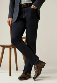 Dunkelblaue Jeans mit kontrastierenden gelben Nähten, kombiniert mit braunen Leder-Brogues, vor einem Hintergrund aus einem Holzhocker.