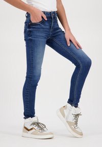 Úzké džíny v tmavě modrém denimu s pěti kapsami, lehkým vyblednutím a zipem. Spojené s bílými a béžovými teniskami na vysoké podrážce.