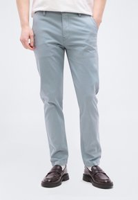 Άνδρας που φοράει ανοιχτό μπλε παντελόνι στενής εφαρμογής και σκούρα καφέ δερμάτινα loafers, στέκεται με τα χέρια στις τσέπες μπροστά σε απλό φόντο.