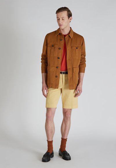 Chaqueta marrón de botones con cuatro bolsillos sobre una camisa roja, combinada con pantalones cortos amarillos y calcetines marrones, llevada con mocasines negros.