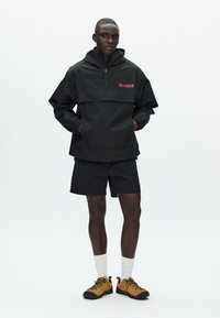 Čierna mikina na zapínanie na štvrťzips, s elastickými manžetami a červeným logom „Double V“. Nosená s čiernymi šortkami a hnedými topánkami.