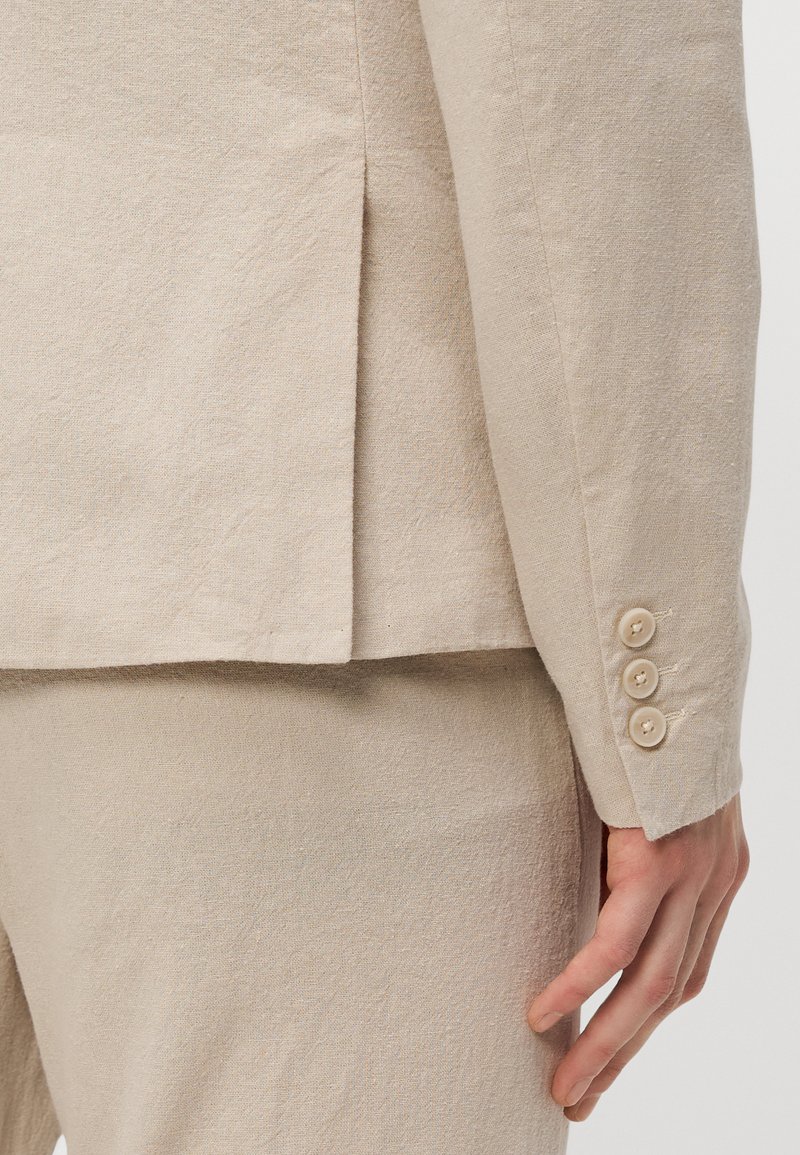 Veste de costume en lin beige avec un revers cranté, dotée d'une fente au dos et de quatre boutons clairs sur les poignets. La texture est légèrement froissée.