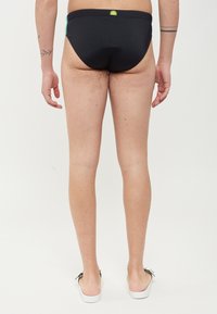 Slip da bagno neri con logo verde sulla parte posteriore, caratterizzati da una vestibilità bassa e un tessuto morbido e elasticizzato, abbinati a scarpe bianche senza lacci.