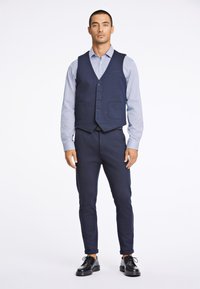 Man staat, draagt een marineblauw vest en broek met een lichtblauw gestreept overhemd en zwarte nette schoenen op een witte achtergrond.