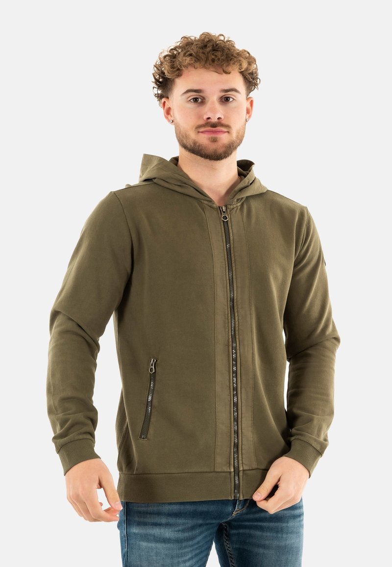 Sweat à capuche zippé vert olive avec une fermeture éclair à l'avant, poches latérales avec fermetures éclair et poignets côtelés. Tissu doux au toucher lisse.