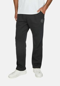 Black sweatpants in soft fabric, featuring a printed logo on the thigh, side pockets, and a relaxed fit. Paired with white sneakers.