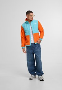 Giacca in pile con blocchi di colore turchese e arancione, con chiusura a zip, due tasche frontali e polsini a coste. Il modello indossa jeans blu.