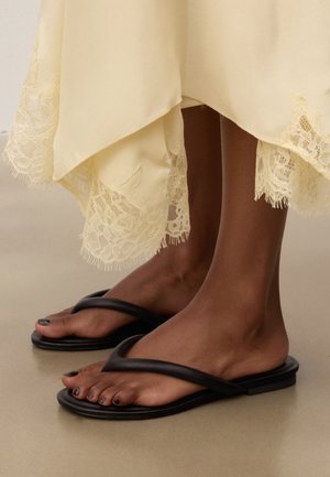 Füße in schwarzen Zehentrennern, kombiniert mit einem cremefarbenen Kleid mit Spitzenbesatz, stehend auf einem glatten beigen Boden.