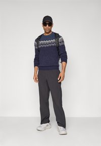 Man in een gedessineerde marineblauwe sweater, zwarte broek, witte sneakers, zwarte pet, donkere zonnebril en een zwarte rugzak staat tegen een effen grijze achtergrond.