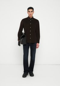 Brun kordfløyels skjorte med knapper, slim-fit mørke denim jeans, svart lærveske og svarte sko, satt mot en enkel hvit bakgrunn.