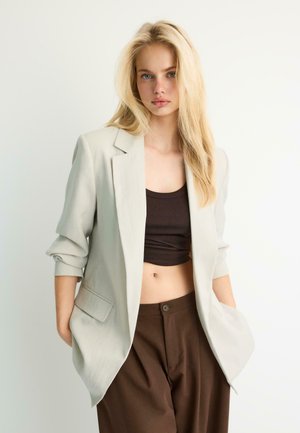Ung kvinde med langt blondt hår iført en lys beige blazer, mørkebrunt crop top og matchende brune bukser, stående med hænderne i lommerne.