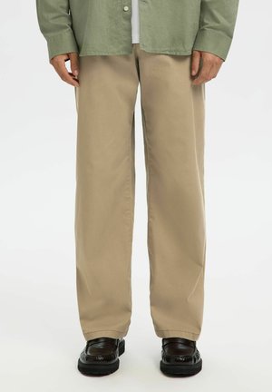 Homme portant un pantalon beige droit, des mocassins noirs et une chemise verte à boutons, debout sur un fond blanc.
