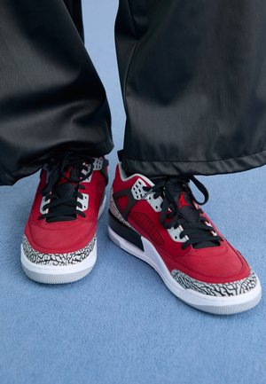 Personne portant un pantalon noir et des baskets à motifs rouges, noirs et gris, debout sur un tapis bleu clair.