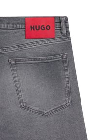 Vaqueros de mezclilla gris con una etiqueta rectangular roja que dice "HUGO". Presentan un bolsillo trasero y costuras sutiles que enfatizan un diseño elegante.