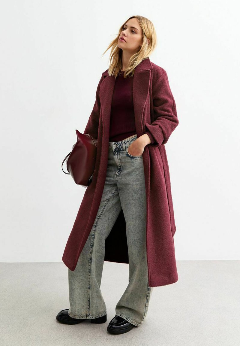 Cappotto di lana bordeaux con taglio dritto e ampie alette, abbinato a un top rosso intenso e jeans a gamba larga grigio chiaro. Scarpe di pelle nere.