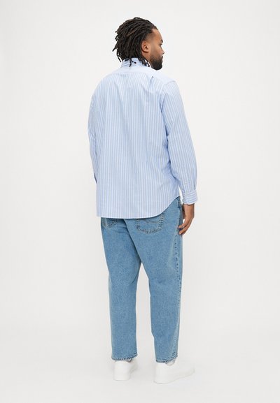 Camisa azul-clara às riscas com mangas compridas e bainha arredondada, combinada com jeans de ganga clara de corte descontraído e ténis brancos.