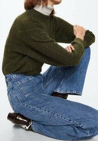 Jersey de color verde oliva con cuello alto, combinado con pantalones vaqueros de mezclilla de corte acampanado y cintura alta en un tono azul claro. Botines brillantes marrones.