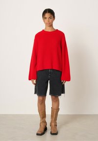 Pull en maille côtelée rouge avec des épaules tombantes, associé à un short en denim noir. Bottes marron jusqu'au mollet avec des boucles et une semelle épaisse.